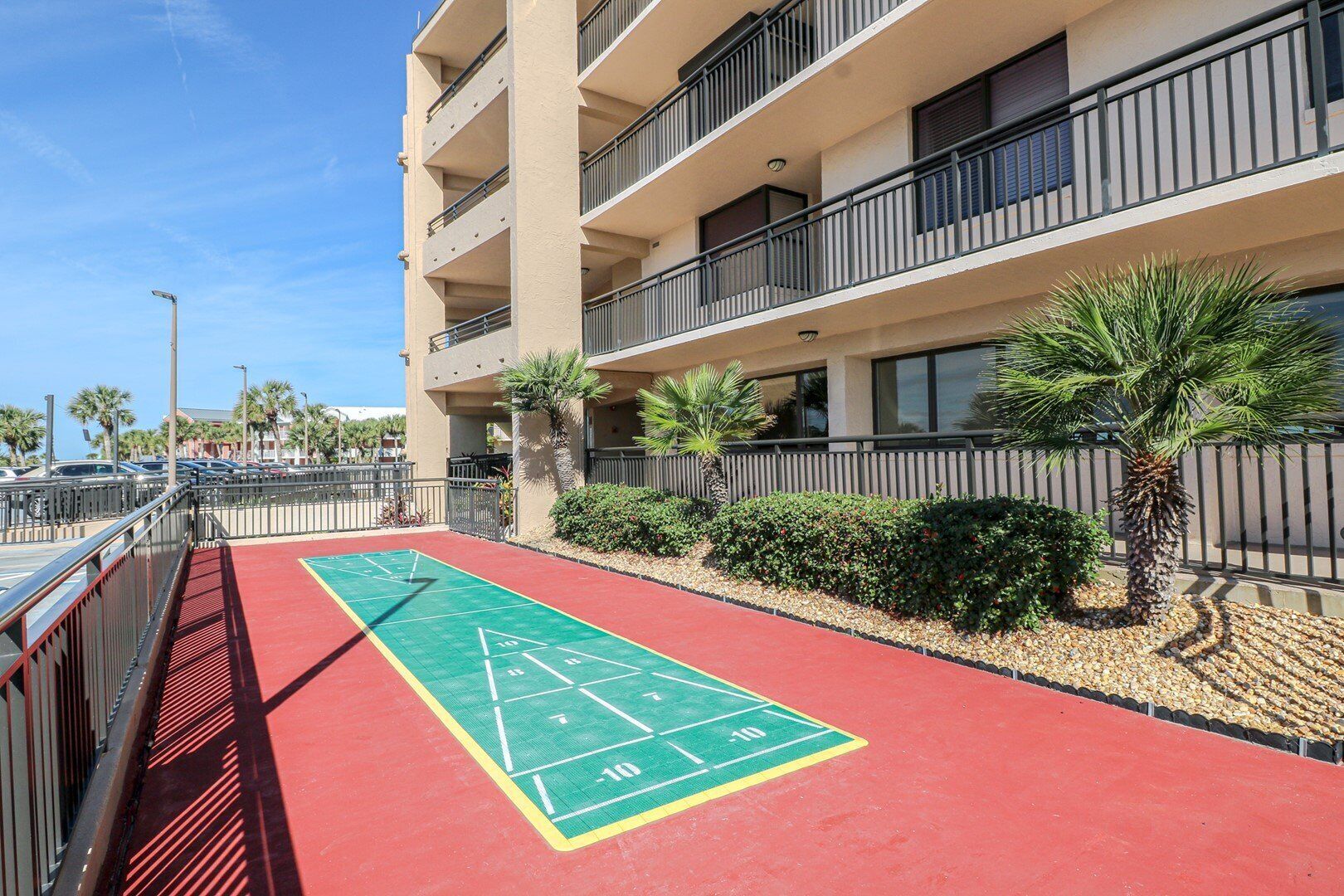 A multi-story building with balconies, a red and green shuffleboard court, palm trees, and a parking area.