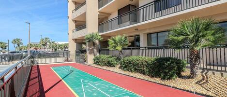 A multi-story building with balconies, a red and green shuffleboard court, palm trees, and a parking area.