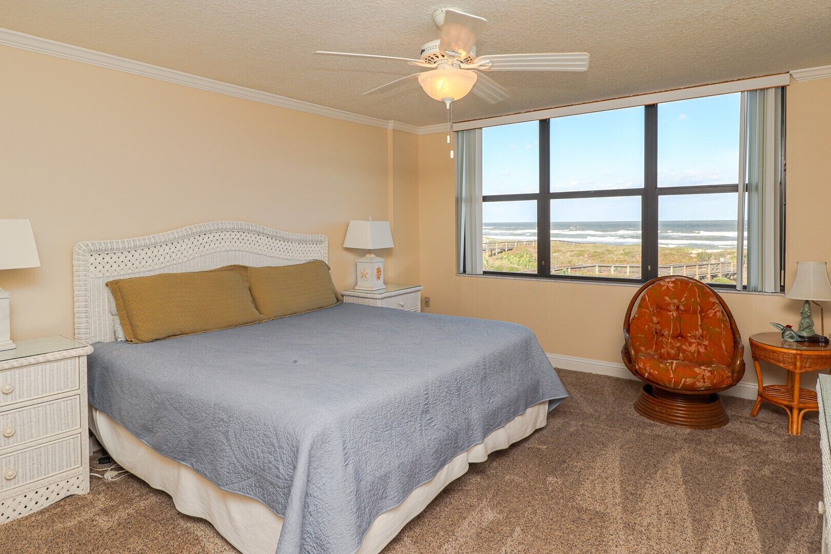 A bedroom with a large bed, a ceiling fan, a view of the ocean, and a wicker chair.