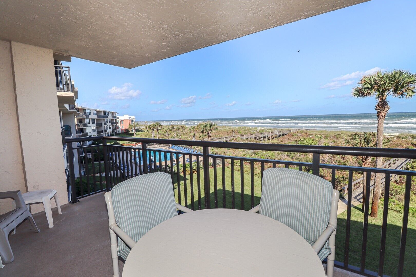 Un balcone con due sedie a righe e un tavolo, con vista su una spiaggia e una passerella in legno, con palme.