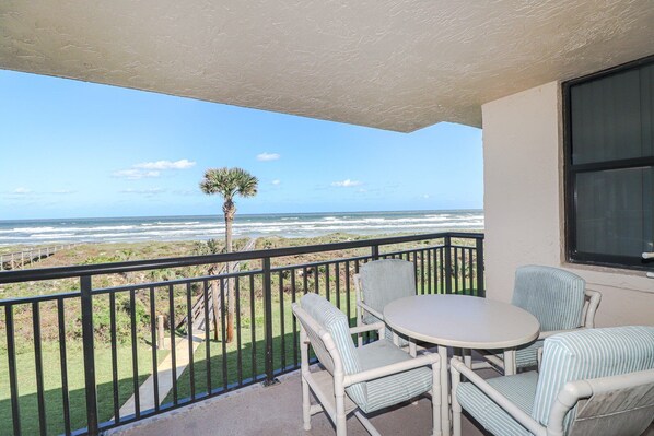 A balcony with a view of the ocean, a palm tree, and a table with striped chairs.