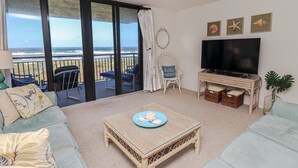 Una sala de estar con un televisor grande, una mesa de centro de mimbre y vistas al océano a través de amplios ventanales.