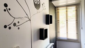 Une pièce dont un mur est décoré d’une fresque représentant un arbre et un hibou, avec un radiateur et deux haut-parleurs noirs fixés au mur.