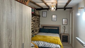 Une chambre avec un mur en pierre, un lit recouvert d’une couverture jaune, une table de chevet en bois et un luminaire suspendu.