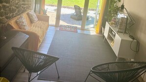 Un salon chaleureux avec un mur en pierre, un canapé agrémenté de coussins à motifs floraux, une table basse et deux chaises noires. On y trouve également un meuble blanc sur lequel sont posés une plante et une lampe.