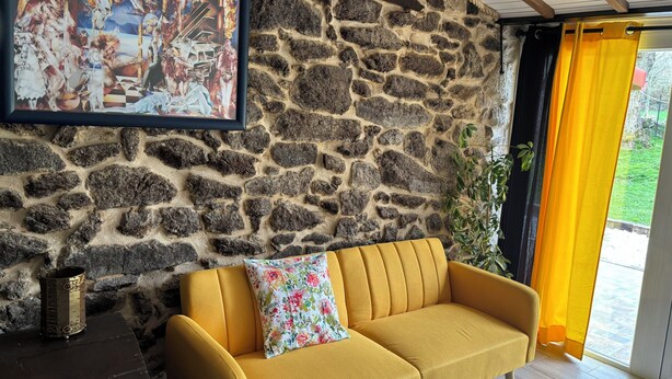 Une pièce avec un mur en pierre, un tableau encadré, un canapé jaune orné d’un coussin à motifs floraux et une fenêtre avec des rideaux jaunes.