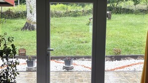 Une porte-fenêtre coulissante donne sur un espace patio extérieur doté d’un sol carrelé en pierre, de plantes en pot et d’une pelouse.