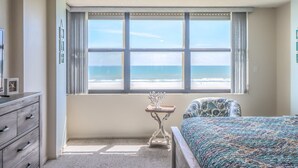 Een slaapkamer met een bed, een stoel, een kapstok, een tafeltje en een raam met uitzicht op de oceaan.