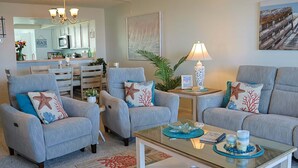 Een zithoek met twee grijze fauteuils, een glazen salontafel, een lamp en een keuken op de achtergrond.