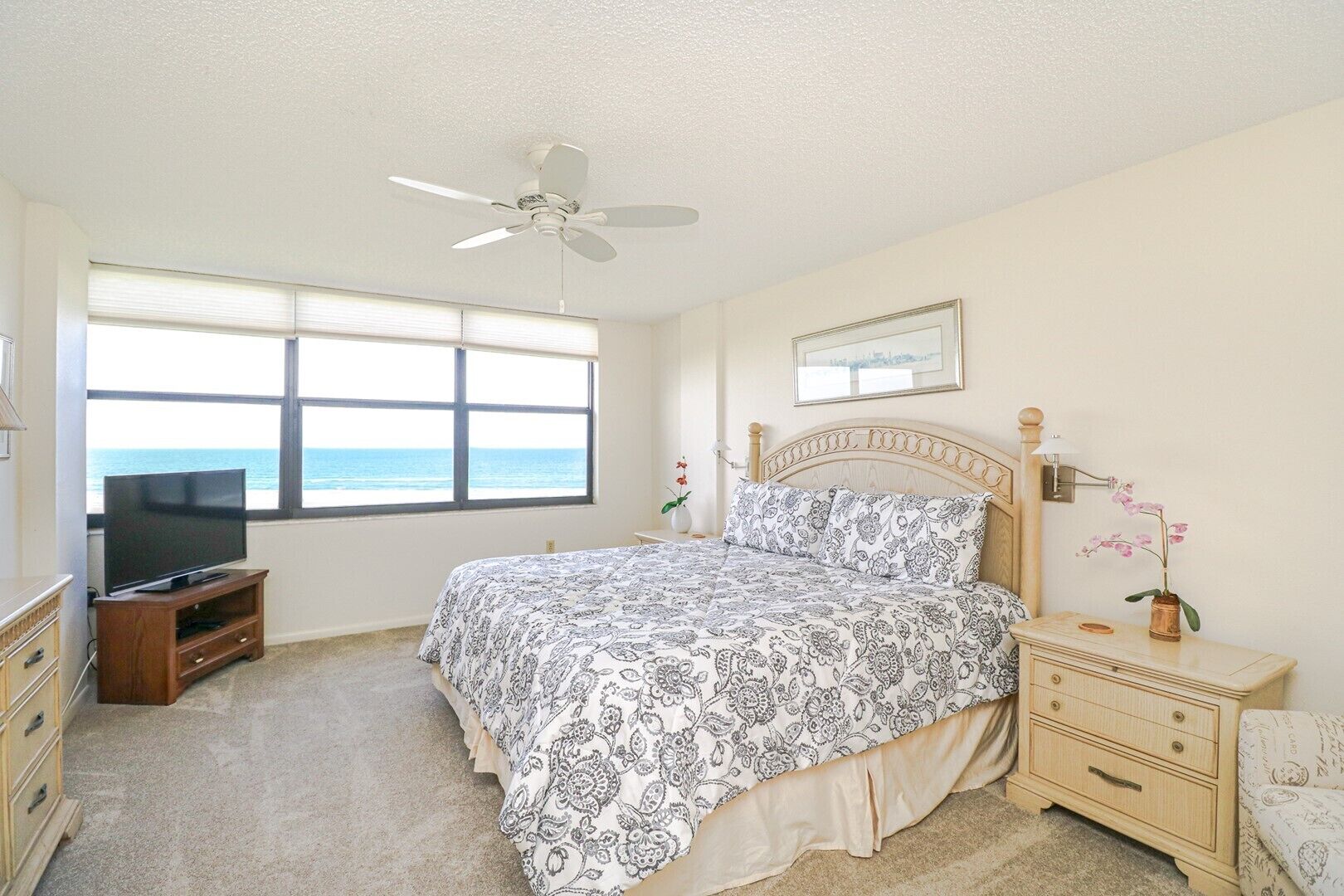 A bedroom with a bed, a TV, a nightstand, a dresser, a ceiling fan, and a window with a view of the ocean.