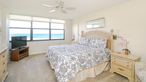 Ein Schlafzimmer mit Bett, Fernseher, Nachttisch, Kommode, Deckenventilator und einem Fenster mit Blick auf den Ozean.