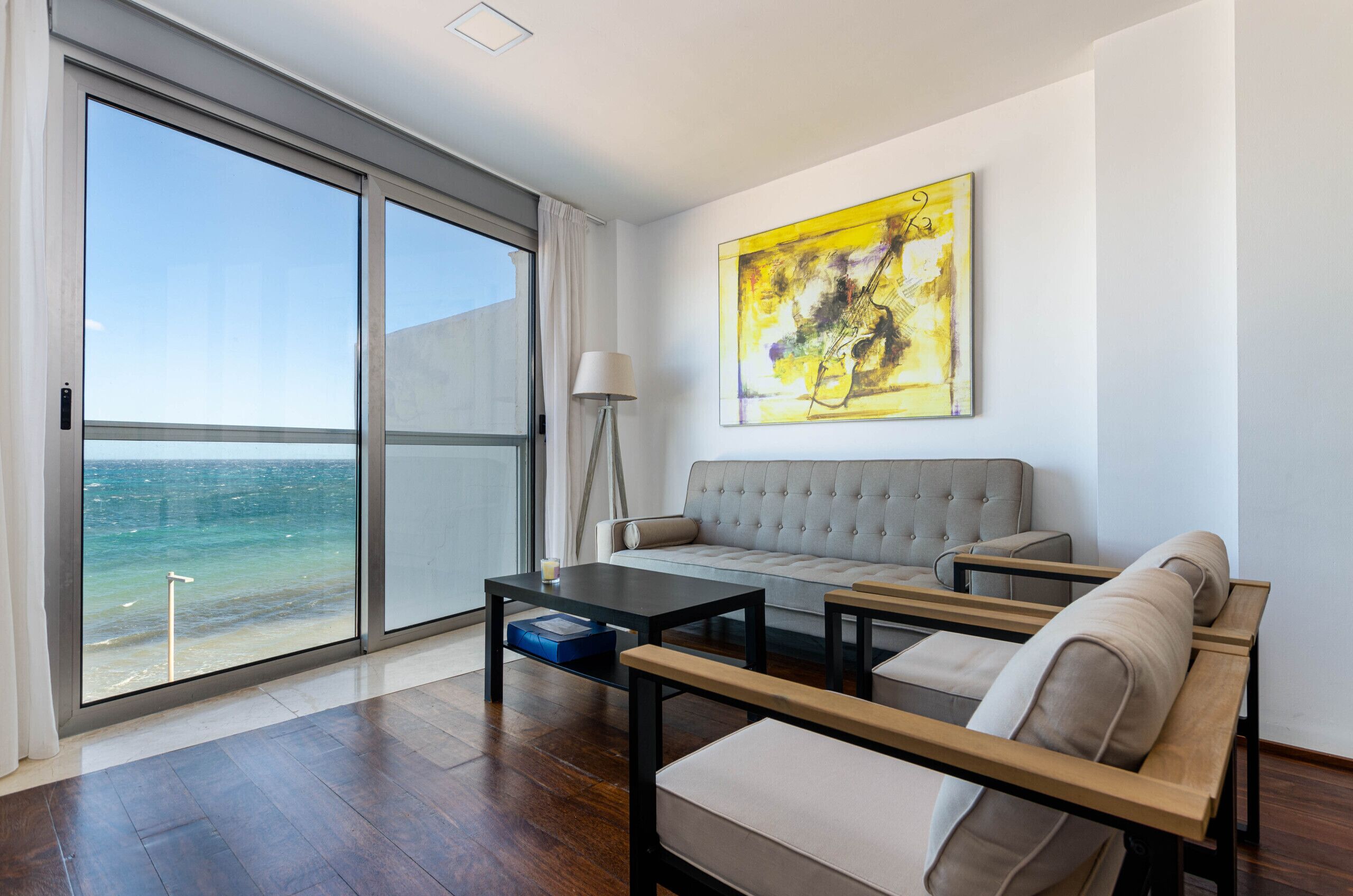 A modern living room with a sofa, two armchairs, a coffee table, and a floor lamp. A large window offers a view of the sea.