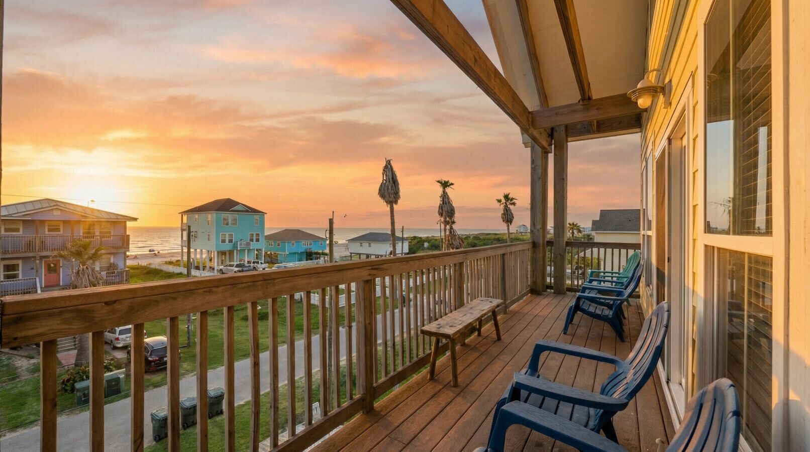 A wooden deck with a bench and chairs, overlooking a beach and ocean during sunset.