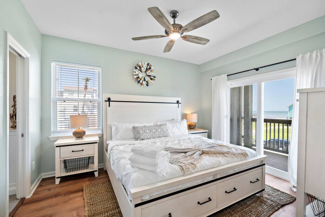 A bedroom with a bed, nightstands, a ceiling fan, a decorative wall piece, and a view of the outdoors.