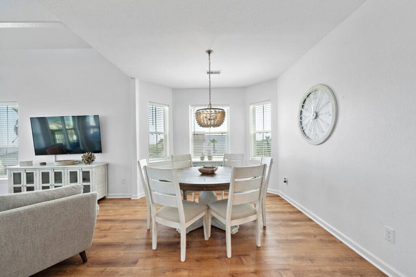 A modern living room with a gray couch, a flat-screen TV, and a dining area with a round table and white chairs.
