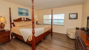 A four-poster bed with a wooden finish, a nightstand with a lamp, a dresser with a mirror, and a window with blinds. - Creston House 11C~Watch the Waves Roll in at this Oceanfront Beauty! (St. Augustine)