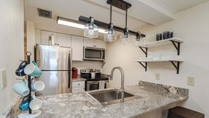 A kitchen with a granite countertop, stainless steel appliances, a hanging light fixture, and a bar stool. - Creston House 11C~Watch the Waves Roll in at this Oceanfront Beauty! (St. Augustine)