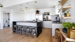 Cocina moderna con armarios blancos, una isla negra y tres taburetes. Hay lámparas colgantes y estantes con plantas y utensilios de cocina.
