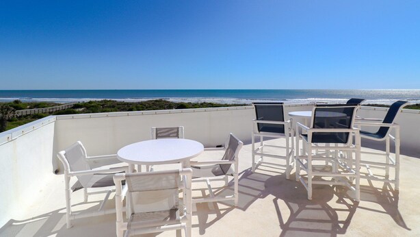 Una terraza con mesa y sillas blancas, con vistas a una playa y al océano.
