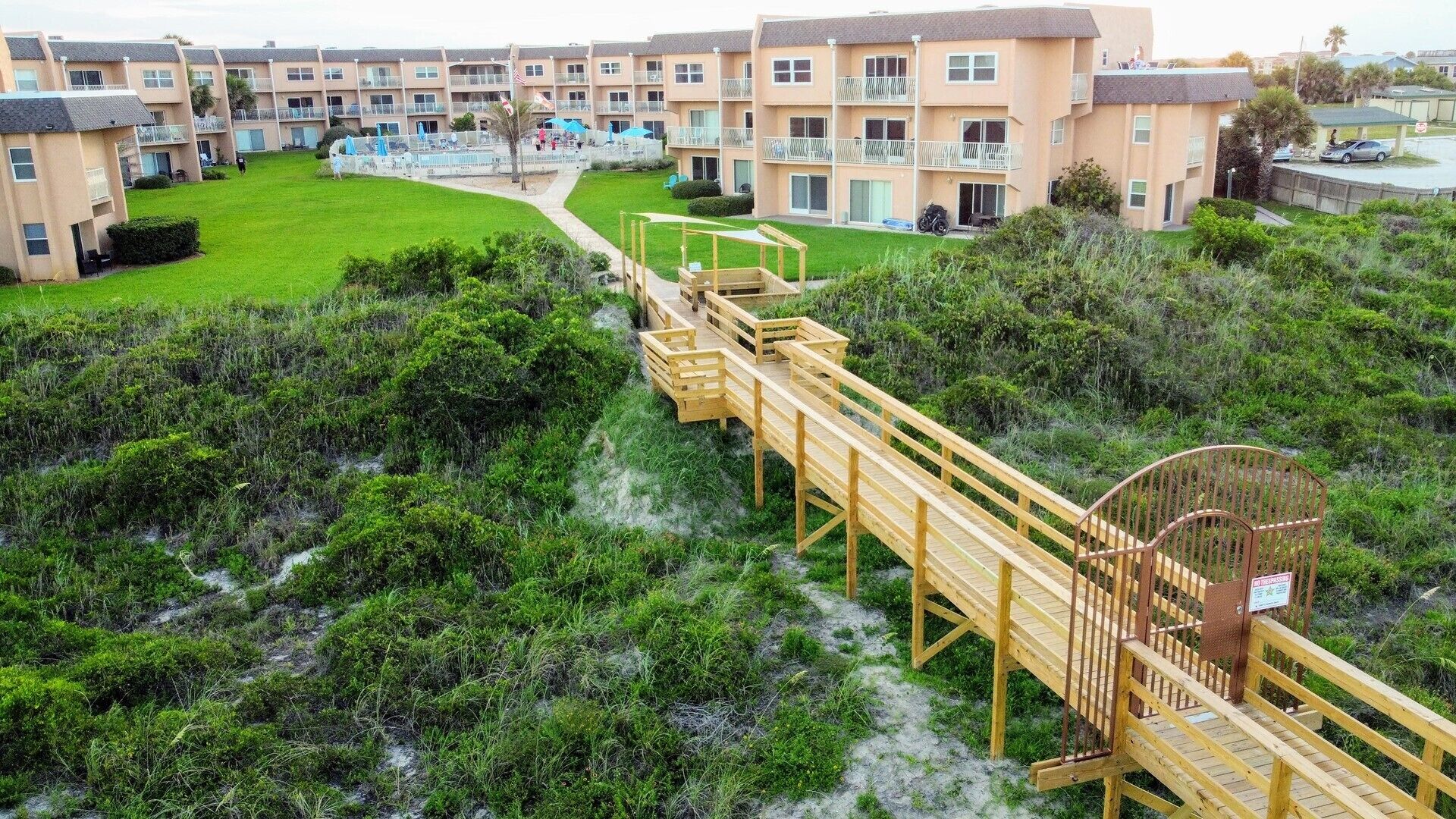 A beachfront hotel with multiple-story buildings, a wooden boardwalk leading to the beach, and a sandy path.