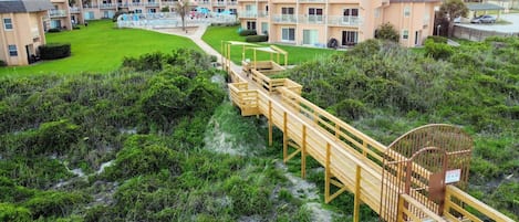 A beachfront hotel with multiple-story buildings, a wooden boardwalk leading to the beach, and a sandy path.