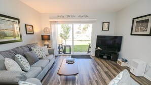 A living room with a grey sofa, a wooden coffee table, a TV, and a large window with a view of the outdoors. - Enjoy this one bedroom condo overlooking the pool, Creston House 5A (St. Augustine)