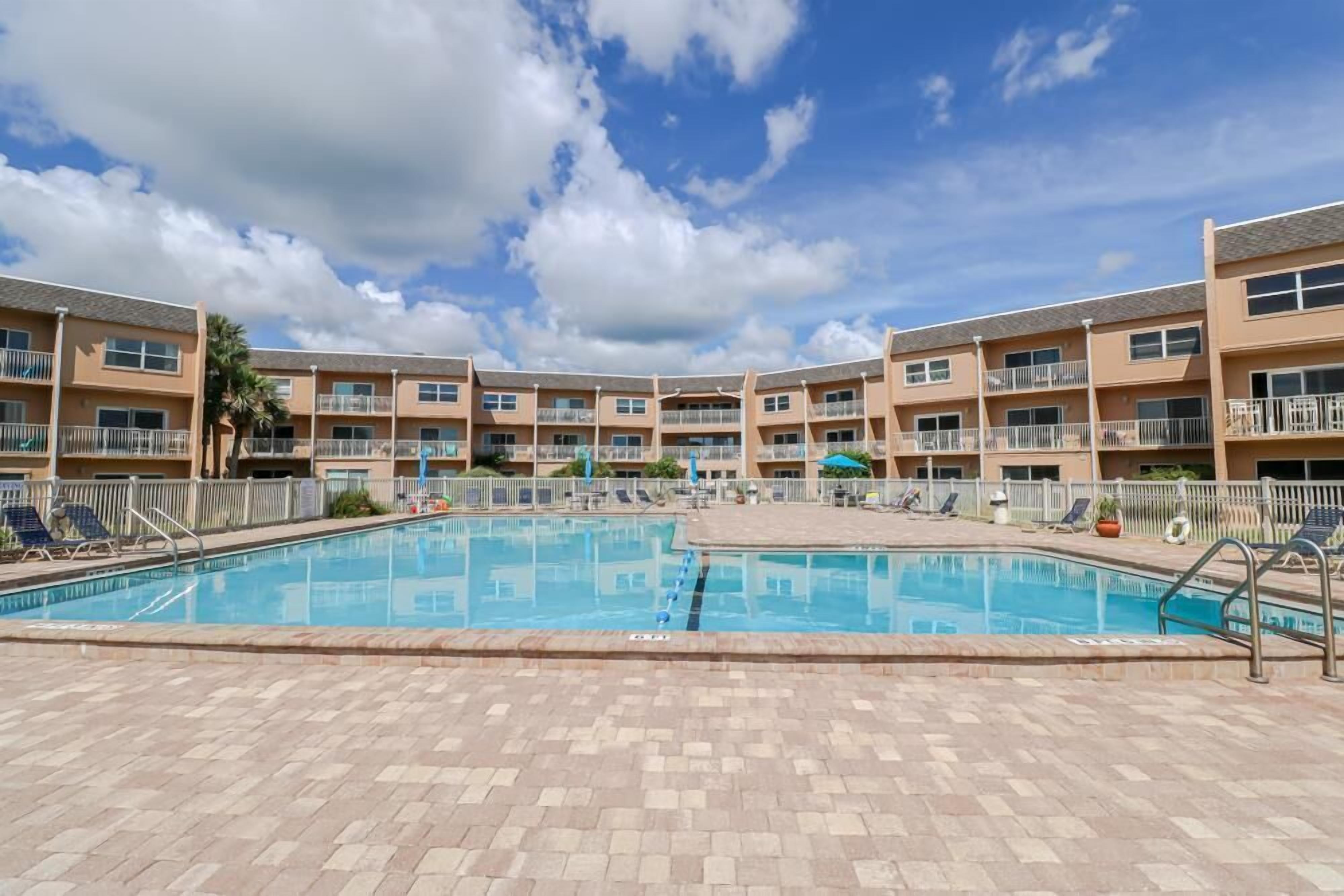A swimming pool surrounded by a multi-story building with balconies, a clear sky, and a paved deck area.