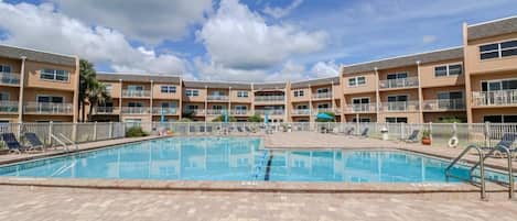 A swimming pool surrounded by a multi-story building with balconies, a clear sky, and a paved deck area.