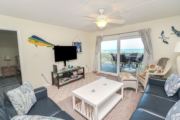 A living room with a flat-screen TV, a ceiling fan, a bookshelf, and a view of the ocean through a sliding glass door.