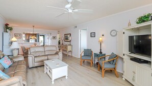 A living room with a sofa, chairs, a coffee table, a TV, and a ceiling fan. - Island South 33 Ocean View Condo On Third Floor (St. Augustine)
