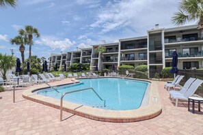 Piscina com espreguiçadeiras e guarda-sóis, rodeada por um edifício de vários andares com varandas.