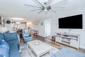 A modern living room with a ceiling fan, a flat-screen TV, a sofa, a coffee table, and a dining area in the background.