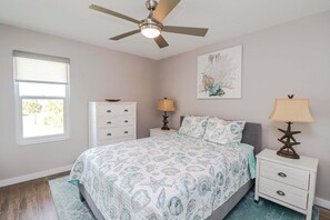 A bedroom with a bed, a ceiling fan, a nightstand with a lamp, a dresser, and a window.