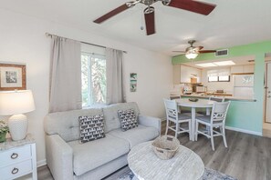 A living room with a sofa, dining table, chairs, a ceiling fan, and a kitchen in the background.