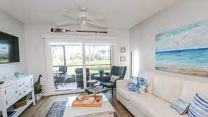 Un salon spacieux avec un canapé blanc, une table basse et un tableau représentant une plage. Un téléviseur est fixé au mur et un ventilateur de plafond est installé.