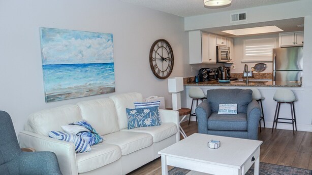 Un salon avec un canapé blanc, un fauteuil bleu et une table basse. Un tableau représentant une scène de plage est accroché au mur.