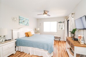 A bedroom with a bed, nightstands, a desk, a ceiling fan, a window, and artwork on the walls.