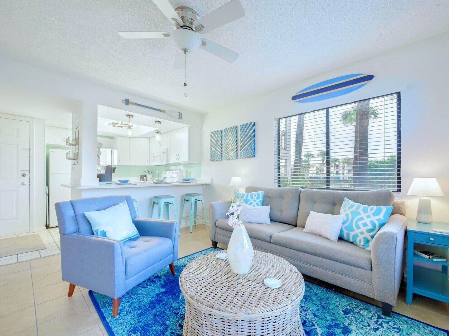 A modern living room with a grey sofa, blue armchair, and a round wicker coffee table. There is a kitchen area with white cabinets and a large window with blinds.