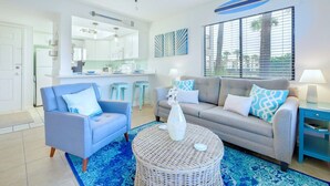 Una sala moderna con un sofá gris, un sillón azul y una mesa de centro redonda de mimbre. Hay una zona de cocina con armarios blancos y un ventanal con persianas.