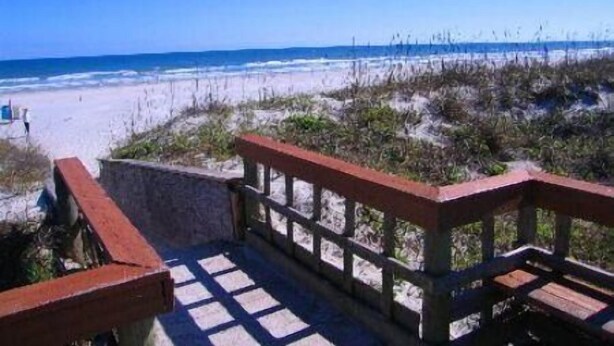 Una terraza de madera con un muro de piedra que da a una playa de arena y vegetación escasa.