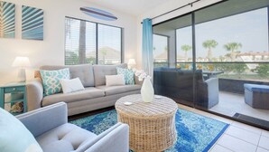Un salón moderno con un sofá gris, cojines azules estampados, una mesa de centro de mimbre y vistas al exterior a través de amplios ventanales.