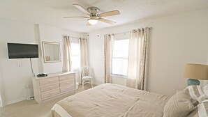 Une chambre à coucher avec un lit, une commode, une chaise, un ventilateur de plafond et un téléviseur fixé au mur.
