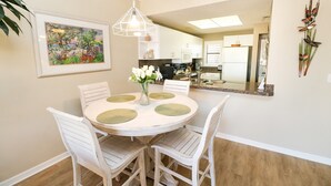 Un comedor con una mesa redonda, sillas blancas y un jarrón de flores. Hay un cuadro en la pared y se ve una cocina al fondo.
