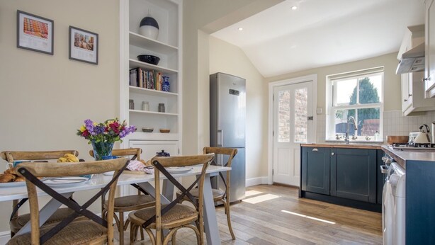 A modern kitchen with a dining area, wooden chairs, a white table, and a built-in bookshelf.