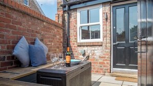 A patio area with a wooden bench, a small table with a bottle of wine and glasses, and a brick wall.