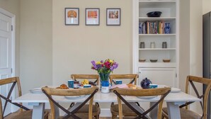 A dining area with a white table, wooden chairs, a vase of flowers, and a bookshelf with books and decorative items.