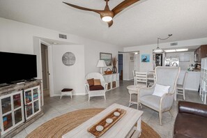 A living room with a television, white furniture, a ceiling fan, and a kitchen in the background.