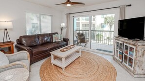 A living room with a brown leather sofa, a white coffee table, a wicker chair, a television on a wooden cabinet, and a ceiling fan. - Ocean Village Club Condo G22 (St. Augustine)