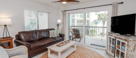 A living room with a brown leather sofa, a white coffee table, a wicker chair, a television on a wooden cabinet, and a ceiling fan.