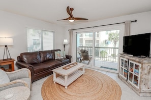 A living room with a brown leather sofa, a white coffee table, a wicker chair, a television on a wooden cabinet, and a ceiling fan.
